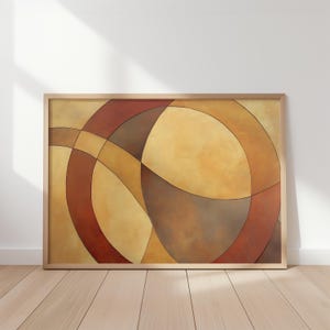 Op de afbeelding: Abstracte kunstafdruk in een houten lijst. Het kunstwerk toont overlappende gebogen vormen in tinten bruin, roest en goud tegen een lichtgele achtergrond. Het stuk wordt tentoongesteld op een witte muur met natuurlijk licht.