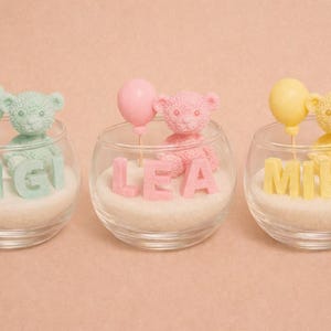 Peut inclure: Trois bols en verre contenant chacun un ours en peluche, un nom et un ballon. Les ours sont vert pastel, rose et jaune. Les noms sont Gigi, Lea et Mika. Les ballons correspondent aux couleurs des ours. Les bols sont sur une surface blanche.