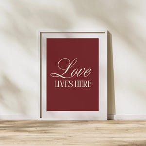 Op de afbeelding: Een ingelijste print met een bordeauxrode achtergrond en de woorden "Love Lives Here" in elegant cursief schrift. De witte lijst contrasteert met het diepe rood, waardoor het een aantrekkelijk stukje wandkunst is.