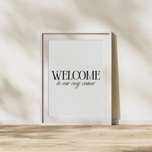 Op de afbeelding: Een witgelijste print met de tekst "WELCOME to our cosy corner" in zwart schrift. De print staat op een houten vloer en tegen een witte muur. De afbeelding heeft een minimalistisch ontwerp, geschikt voor interieurdecoratie.