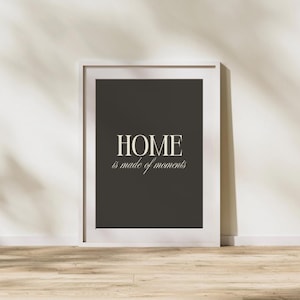 Op de afbeelding: Een ingelijste print met de woorden "HOME is made of moments" in een witte, elegante letter. De achtergrond is donkergrijs en de lijst is wit. De print wordt gepresenteerd tegen een witte muur en een houten vloer.