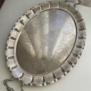 Può includere: Un vassoio da portata ovale color argento caratterizzato da un ornato motivo floreale inciso al centro e da un bordo decorativo smerlato. Il vassoio include due manici in metallo curvi e dettagliati fissati alle estremità opposte.