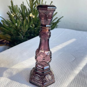 May include: A tall, amethyst-coloured glass candlestick holder with ornate, embossed detailing. The candlestick has a square base and a flared top, with decorative rings and patterns. The glass has a glossy finish. The height is approximately 20 cm.