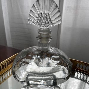 Può includere: Un flacone di profumo in vetro trasparente con un tappo decorativo a forma di ventaglio. La bottiglia ha un corpo arrotondato e poggia su una superficie a specchio. Il tappo presenta un design a taglio diamante.