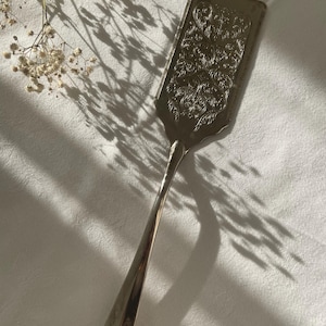 Könnte beinhalten: Silberfarbener Kuchenheber mit einem verzierten, gravierten Blumendesign auf der Klinge. Der Heber hat einen langen, schlanken Griff und liegt auf einer weißen Tischdecke. Das Bild wird von Sonnenlicht beleuchtet, das Schatten wirft. Der Heber ist etwa 25 cm lang.
