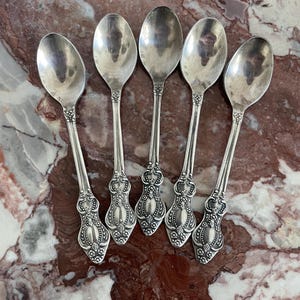 Peut inclure: Cinq cuillères en argent antiques disposées sur une surface en marbre. Les cuillères ont des manches ornés et détaillés avec des motifs floraux et enroulés. Les cuillères reflètent l'environnement. Un ajout classique à toute ménagère.