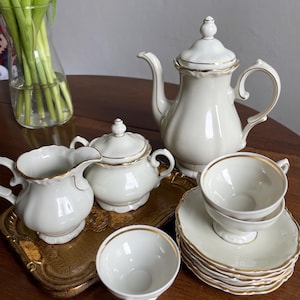 May include: A cream-coloured tea set with gold trim, including a teapot, sugar bowl, creamer, cups, and saucers. The set is arranged on a gold-coloured tray and a wooden table. A vase of green tulips is in the background.