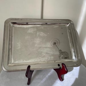 May include: A rectangular silver-toned serving tray with a smooth, reflective surface and a raised, multi-layered border. The tray is positioned on a dark reddish-brown display stand, showcasing its metallic finish and clean, simple design.