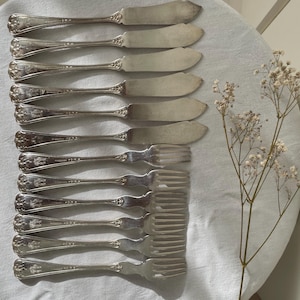 May include: A set of six silver-toned butter knives and six silver-toned forks, each featuring an ornate, embossed floral pattern on the handle. The cutlery is arranged in two parallel rows, with the knives positioned above the forks.