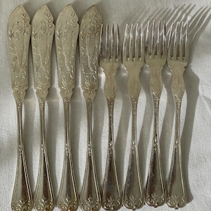 Pode incluir: Um conjunto de oito talheres de metal prateado composto por quatro facas de peixe e quatro garfos. Cada faca de peixe apresenta um design em relevo de peixe e folhagem no cabo. Os garfos têm um padrão decorativo correspondente na base.