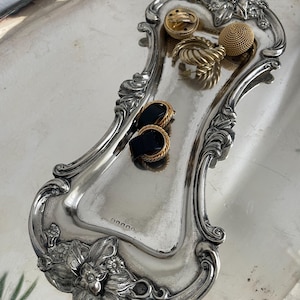Può includere: Una collezione di orecchini color oro, tra cui un paio con centri rettangolari neri e bordi dorati intrecciati, poggiati su un vassoio in metallo argentato, allungato e decorato con dettagli floreali e marchi visibili.