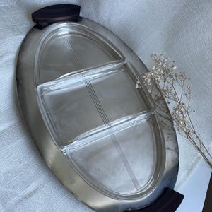 Könnte beinhalten: Ein ovales Serviertablett mit einem polierten, silberfarbenen Metallrahmen und dunkelbraunen Holzgriffen an beiden Enden. In der Mitte befindet sich ein klarer Glaseinsatz, der in drei Abschnitte unterteilt ist, die jeweils mit feinen Linien verziert sind.
