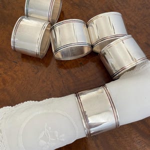May include: A collection of six silver napkin rings with a polished finish. Each ring has a thin, colored band near the top and bottom edges. One ring holds a white, embroidered napkin, showcasing a classic table setting.