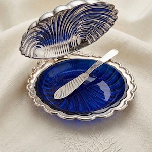 May include: A silver-plated serving dish with a deep blue glass interior, shaped like a clam shell. A matching silver knife rests inside. The dish sits on a cream-coloured cloth with a starfish and coral design.