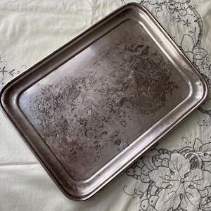 Puede incluir: Una bandeja rectangular de plata antigua con una superficie desgastada y texturizada. La bandeja tiene un borde ligeramente elevado y está colocada sobre una tela crema con un estampado floral.