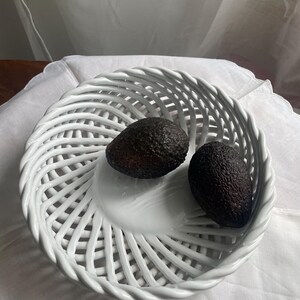 Op de afbeelding: Een witte keramische mand met een geweven ontwerp bevat twee rijpe avocado's. De mand staat op een witte doek, wat een strakke esthetiek creëert. De avocado's hebben een donkere, getextureerde schil.
