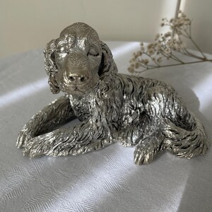 Könnte beinhalten: Eine metallisch silberfarbene Figur eines Spaniels in liegender Position. Die Skulptur zeichnet sich durch detailliertes, strukturiertes Fell am ganzen Körper, lange Schlappohren und einen ruhigen Gesichtsausdruck mit geschlossenen Augen aus.