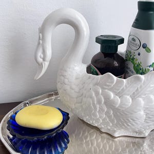 以下が含まれることがあります： 白鳥の形をした白い陶器製の容器には、ダークブラウンのボトルと緑のアクセントが入った白いボトルの2本が入っており、銀色のトレイの上に置かれています。その隣には、淡い黄色の丸い石鹸が入った、縁が波打ったコバルトブルーのガラス皿があります。