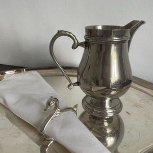 Puede incluir: Una jarra de crema de metal plateado pulido con asa curva y pico vertedor, acompañada de un servilletero de metal plateado con forma de cisne que sostiene una servilleta de tela blanca doblada, todo sobre una bandeja de metal plateado.