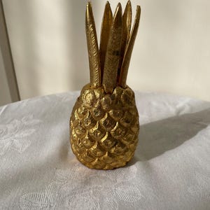 Peut inclure: Une figurine d'ananas de couleur dorée avec un corps texturé à motif de losanges et des feuilles pointues dressées vers le haut. L'objet présente une finition métallique et une base arrondie, se tenant verticalement comme objet décoratif.