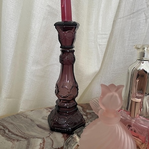 Könnte beinhalten: Ein hoher, dunkelvioletter Kerzenhalter aus Glas mit floralem Design, der eine dunkelrosa Kerze hält. Der Kerzenhalter steht neben einer rosa Parfümflasche und anderen dekorativen Gegenständen.