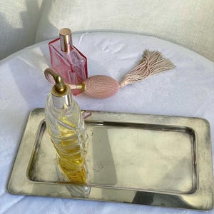 Puede incluir: Una bandeja rectangular de metal plateado que contiene dos frascos de perfume de vidrio. Uno es transparente con un diseño en espiral y líquido amarillo, el otro es rosa con un atomizador de pera cubierto de tela beige y una borla.