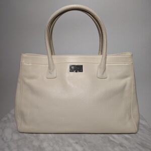 May include: Cream leather tote bag with two rounded handles and a rectangular silver clasp. The bag has a rectangular shape and a textured surface. The handles are cream-coloured and appear to be stitched.