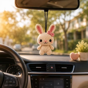 May include: A cream-colored crochet rabbit hanging ornament with long ears, pink inner ear details, a small pink bow, black bead eyes, and a stitched mouth. The plush toy is suspended by a black cord from a vehicle rearview mirror.
