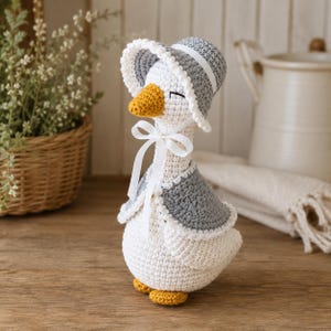 May include: A crochet goose figurine featuring a white body, a yellow beak, and yellow feet. The goose wears a grey bonnet with a white scalloped trim, a matching grey capelet, and a white ribbon tied in a bow around its neck. The eyes are closed.