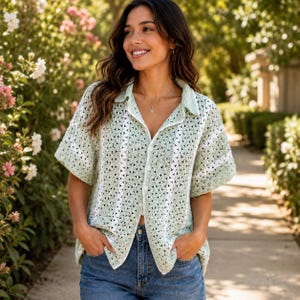 May include: A short-sleeved crochet button-down shirt in a light sage green hue, featuring a repeating floral-inspired pattern and vertical white stripes. The garment has a classic collar, a front button closure, and a relaxed, boxy fit.