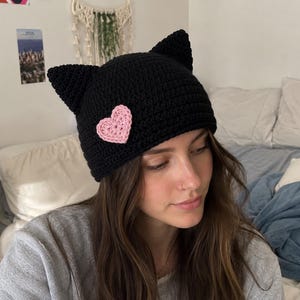 May include: A black crocheted beanie featuring two pointed cat-ear shapes at the top and a small, textured light pink heart appliqué attached to the front side. The hat is constructed with a dense, uniform stitch pattern throughout the fabric.