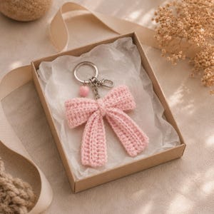 Puede incluir: Un llavero de lazo tejido a ganchillo en color rosa claro, con una anilla de metal plateado, un pequeño cierre de mosquetón y una cuenta redonda de color rosa. El artículo se presenta en una caja de regalo cuadrada de cartón con papel seda.