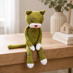 May include: A handmade crochet cat toy featuring a variegated olive green yarn body, white paws, and a white muzzle. The cat has embroidered closed eyes and a small red stitched nose and mouth. The toy is designed to sit upright with a long tail.