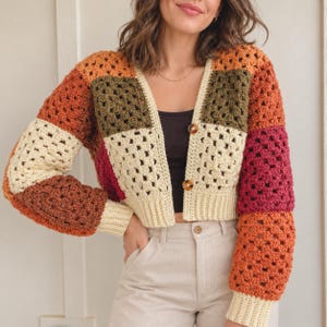 May include: A cropped crochet cardigan featuring a patchwork design of granny squares in shades of cream, olive green, rust orange, and deep burgundy. The garment has long sleeves, a ribbed cream hem and cuffs, and two brown tortoiseshell buttons.