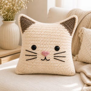 May include: A square-shaped crochet pillow designed to resemble a cat face. The pillow is cream-colored with dark brown triangular ears, two black circular eyes, a small pink nose, and black embroidered whiskers and a mouth on the front.