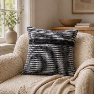 May include: A square throw pillow featuring a black and white textured knit pattern. A horizontal dark gray crocheted band runs across the center, accented with three small, round, black buttons spaced evenly along the length of the band.