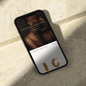 Puede incluir: Un smartphone muestra una p&aacute;gina web de comercio electr&oacute;nico de joyas. La pantalla muestra a una persona con joyas de oro y el texto "Everyday elegance, made to wear". Dos pares de pendientes de aro dorados se muestran en la parte inferior.