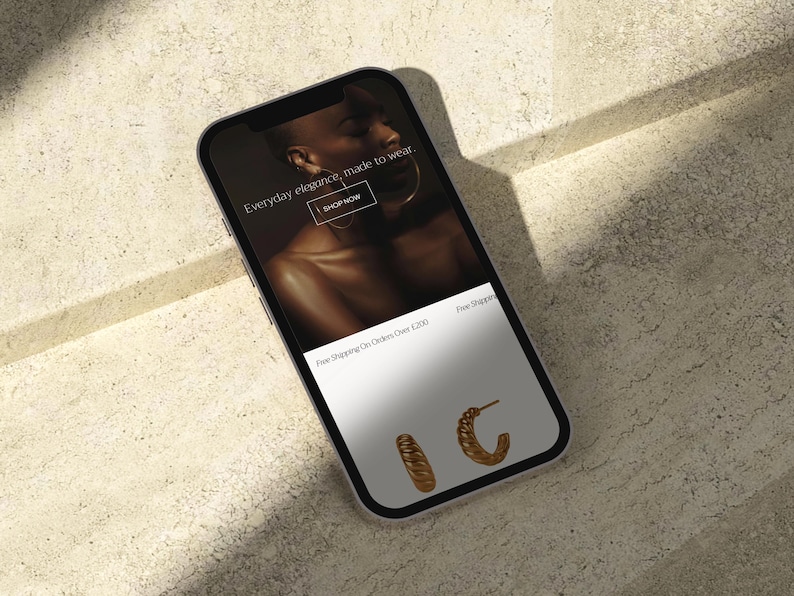 Puede incluir: Un smartphone muestra una p&aacute;gina web de comercio electr&oacute;nico de joyas. La pantalla muestra a una persona con joyas de oro y el texto "Everyday elegance, made to wear". Dos pares de pendientes de aro dorados se muestran en la parte inferior.