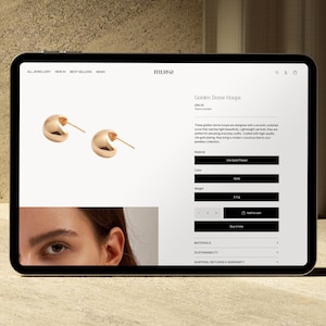 Puede incluir: Una tableta muestra un sitio web de joyer&iacute;a con aros dorados. Los aros son lisos, pulidos y tienen un dise&ntilde;o curvo. El sitio web incluye detalles del producto, como el material, el color y el peso de 0,5 g.