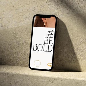 Puede incluir: Un smartphone muestra el texto "#BE BOLD" en su pantalla, con un pendiente dorado y dos anillos dorados. El tel&eacute;fono descansa sobre una superficie clara y texturizada, con sombras.