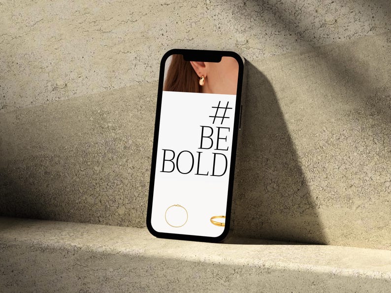 Puede incluir: Un smartphone muestra el texto "#BE BOLD" en su pantalla, con un pendiente dorado y dos anillos dorados. El tel&eacute;fono descansa sobre una superficie clara y texturizada, con sombras.