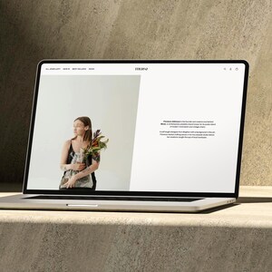 Puede incluir: Un port&aacute;til plateado muestra un sitio web con una mujer sosteniendo flores. El sitio web presenta el logotipo "MUSE" y texto sobre Florence Ellison. El port&aacute;til est&aacute; sobre una superficie clara con un fondo texturizado.