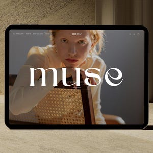 Può includere: Un tablet digitale mostra il sito web di "muse", un marchio di gioielli. Il sito web presenta una donna dai capelli rossi e la parola "muse" in grandi lettere bianche. Il tablet è su una superficie marrone chiaro.