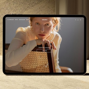 Puede incluir: Una tableta muestra el sitio web de joyas "muse". La imagen presenta a una persona pelirroja, que lleva joyas y se apoya en una silla. El sitio web incluye el texto "Enrich the everyday" y un bot&oacute;n "Shop Now".