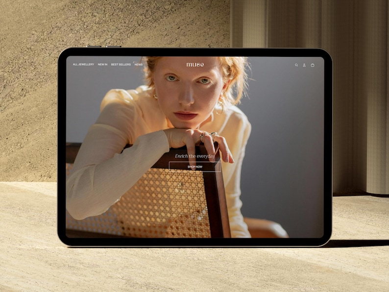 Puede incluir: Una tableta muestra el sitio web de joyas "muse". La imagen presenta a una persona pelirroja, que lleva joyas y se apoya en una silla. El sitio web incluye el texto "Enrich the everyday" y un bot&oacute;n "Shop Now".
