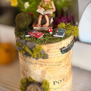 May include: A decorative cylindrical display featuring a small elf figure reading a book while seated on a stack of books. The base is covered in green moss, miniature books titled Gone with the Wind and The Cat in the Hat, and a small arched wooden door.