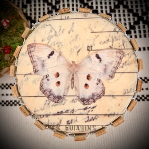 May include: A circular decorative object featuring a cream-colored surface with a printed butterfly graphic and vintage-style cursive script text. The edge is bordered by small, evenly spaced wooden blocks and a thin wire frame structure.