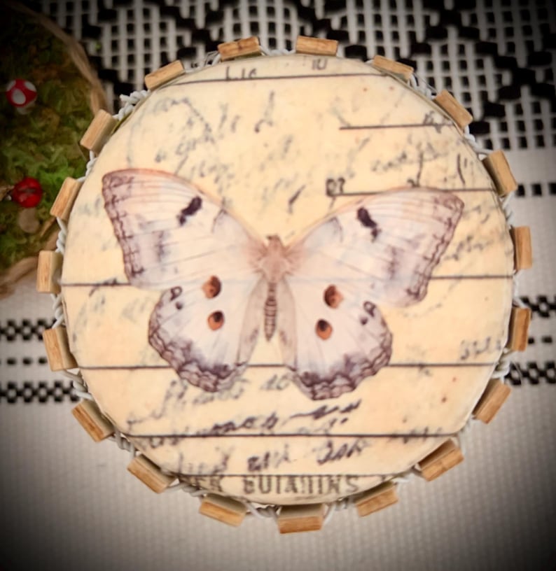 May include: A circular decorative object featuring a cream-colored surface with a printed butterfly graphic and vintage-style cursive script text. The edge is bordered by small, evenly spaced wooden blocks and a thin wire frame structure.