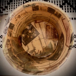May include: A cylindrical container featuring a decoupage design with vintage-style postage stamps, handwritten cursive script, and floral illustrations in shades of beige, brown, and cream. The interior walls and base display a collage of paper.