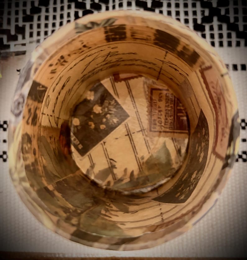 May include: A cylindrical container featuring a decoupage design with vintage-style postage stamps, handwritten cursive script, and floral illustrations in shades of beige, brown, and cream. The interior walls and base display a collage of paper.
